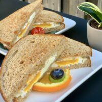 an egg sandwich on toasted bread plated on a white plate with fruit on a black table